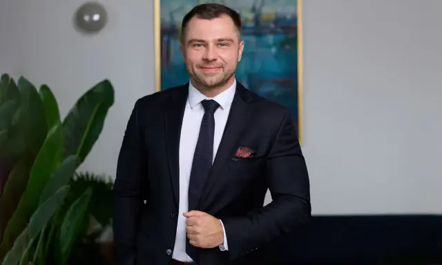 Mężczyzna w garniturze, stojący obok rośliny, z obrazem w tle.