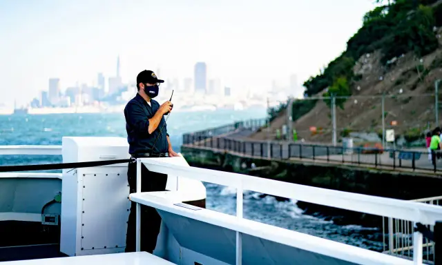 Un homme sur un bateau utilise une radio. Il est en communication, peut-être sur une fréquence vhf libre, avec la ville en arrière-plan.