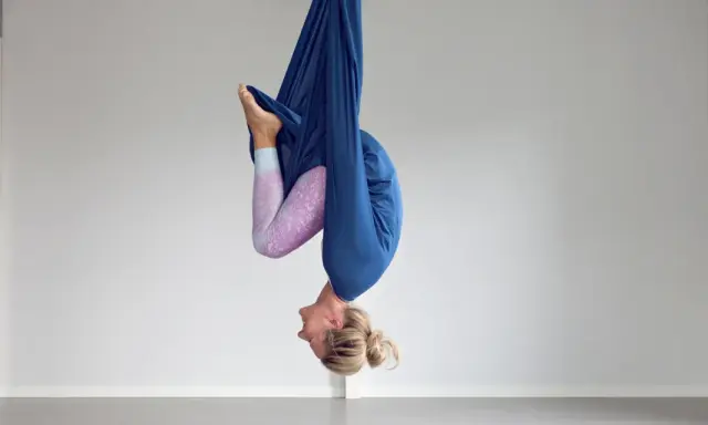 Kvinna hänger upp och ner i en blå tyghängmatta, utövar flying yoga.