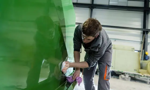 Un ouvrier polit la coque d'un bateau en polyester vert vif dans un atelier.