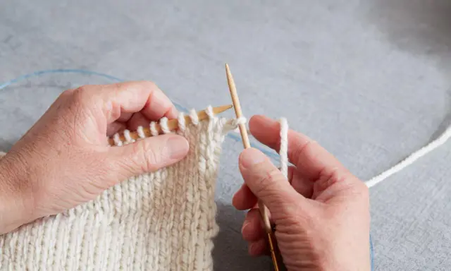 Händer stickar en vit resår med trästickor. En blå kabel syns i bakgrunden.