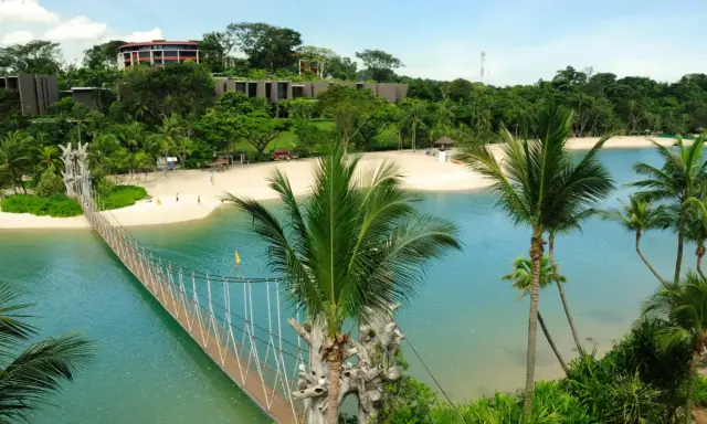 Hängende Brücke über türkisfarbenem Wasser führt zu einem sonnigen singapur strand mit Palmen und Resorts im Hintergrund.