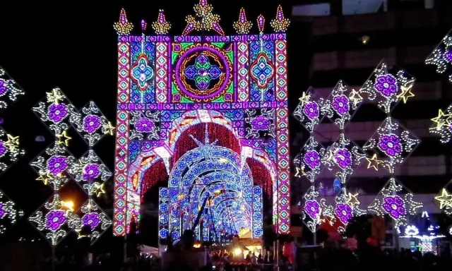Un tunnel di luci colorate e decorazioni luminose, tipiche delle feste patronali, crea un'atmosfera magica e festosa.