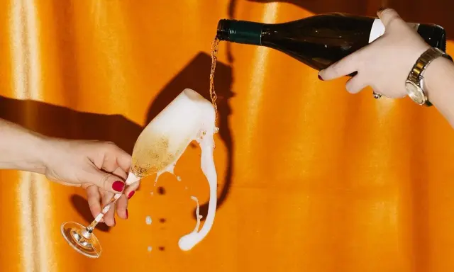 Schaumiges Sektglas läuft über, während eine Flasche auf orangefarbenem Hintergrund eingegossen wird. Eine Szene, die an die **zero dosage definition** erinnert, wo jeder Tropfen zählt.