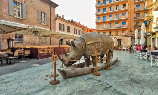 Statua di rinoceronte in una barca, in una piazza di Rimini, dove è nato Fellini. Tavolini all'aperto e palazzi colorati sullo sfondo.
