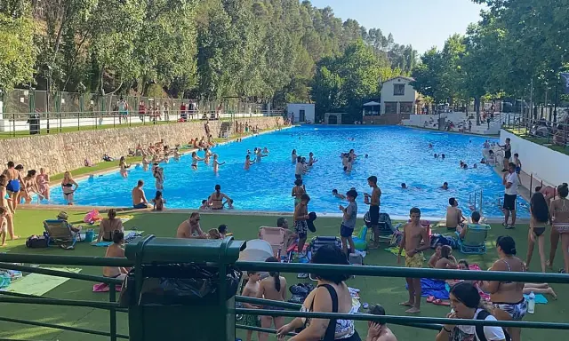 Gente disfrutando de un día soleado en la **piscina segura de la sierra**. Niños y adultos se refrescan en el agua azul, rodeados de árboles y un paisaje montañoso.