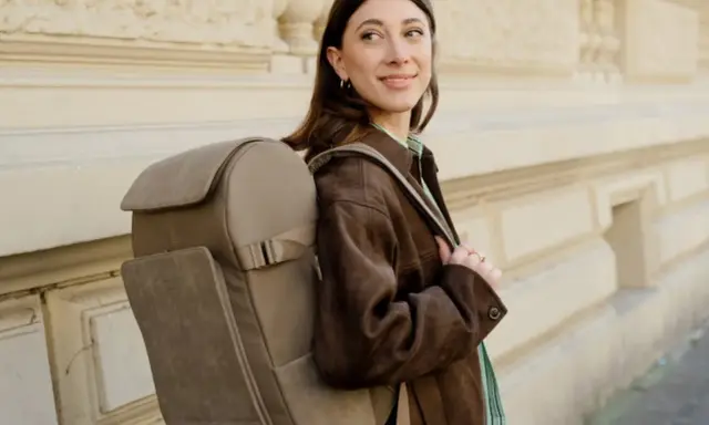A woman smiles, showcasing the latest backpacks trend with a stylish, taupe backpack.