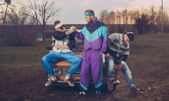 Trzech mężczyzn w dresach i swetrach, uosabiających modę lat 90., siedzi na masce starego samochodu.
