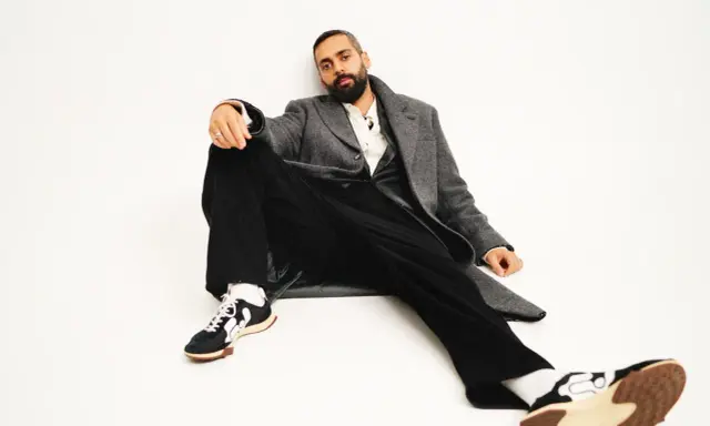 Ardalan Esmaili sitter avslappnat i en grå rock, vit skjorta och svarta byxor. Hans sneakers har en unik design.