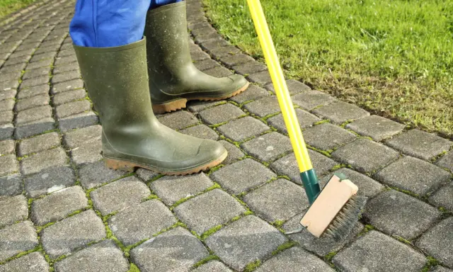 Person en bottes vertes brossant la mousse sur une terrasse pavée. Astuce pour savoir comment enlever la mousse sur une terrasse.