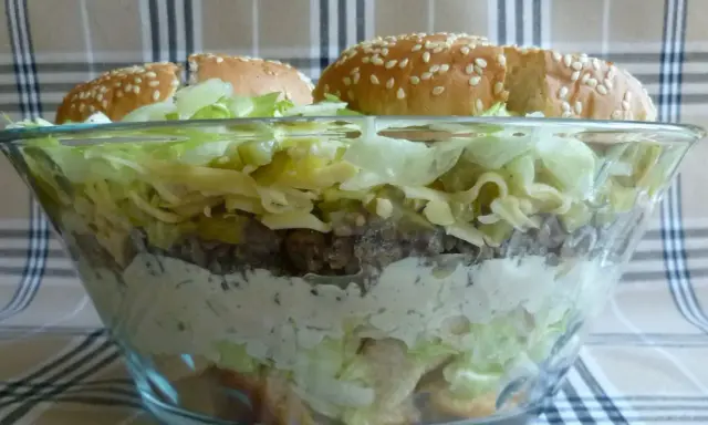 Warstwowa sałatka big mac w szklanej misie, z bułkami na wierzchu.