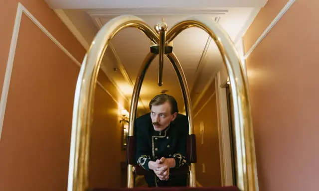 Elegancki hotelarz w historycznym mundurze czeka przy wózku bagażowym w hotelowym korytarzu.