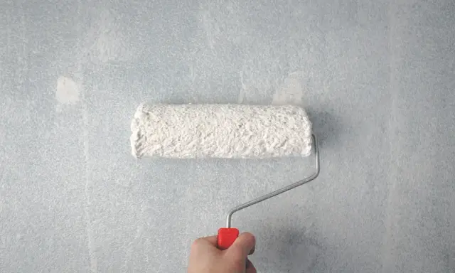 Main tenant, une main tient un rouleau à peinture blanc sur un mur bleu clair. Les traces de rouleau à la lumière révèlent la texture du mur.