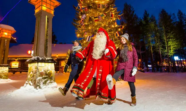 Santa Claus mit zwei Besuchern vor einem Weihnachtsbaum im Rovaniemi Weihnachtsdorf.