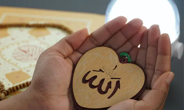 Hände halten ein Herz mit dem Schriftzug "Allah" in arabischer Schrift. Im Hintergrund liegen Gebetskette und ein Buch.