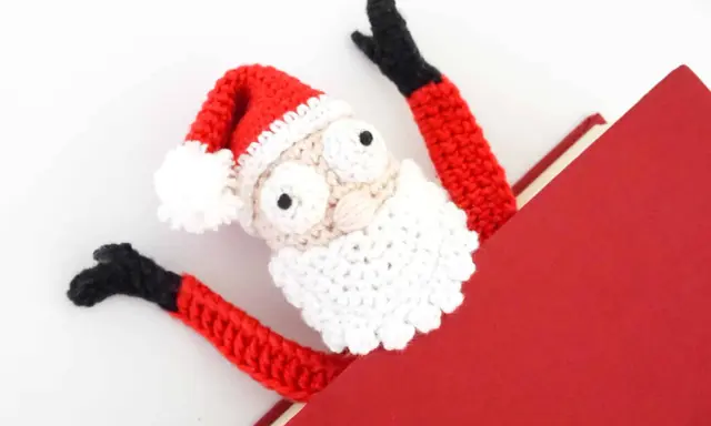 Marcador de libro amigurumi de Papá Noel, con brazos rojos y gorro festivo, asomando de un libro rojo.