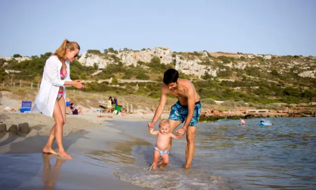 Familia disfrutando de un día en la playa en Menorca con niños. El padre ayuda a su bebé a dar sus primeros pasos en el agua, mientras la madre observa sonriente.