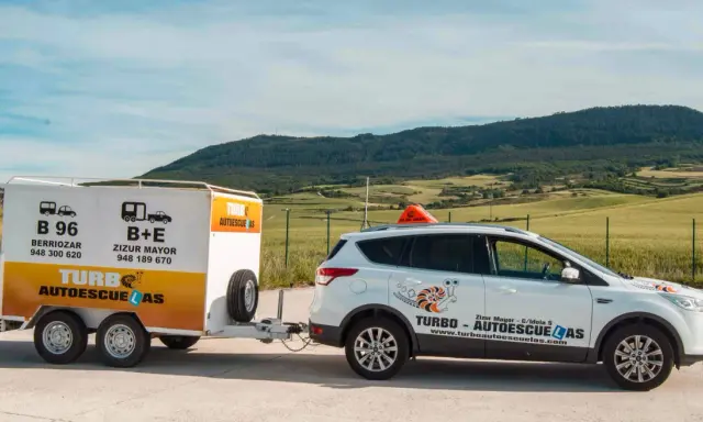 Un coche blanco de **4x4 autoescuela** con remolque, listo para enseñar a conducir en un paisaje rural.