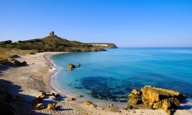 Spiaggia dorata e mare cristallino in un'area marina protetta, con una torre antica su una collina.