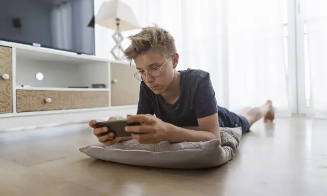 Un adolescent, absorbé par son téléphone, illustre la dépendance aux écrans. Il est allongé sur un coussin, le regard fixé sur l'appareil.
