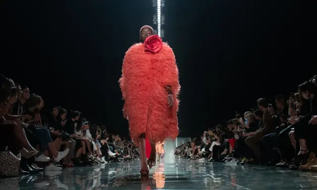 Modella con cappotto vaporoso color corallo e fiore rosso, sfila su una passerella lucida. Il pubblico ammira gli abbinamenti audaci.