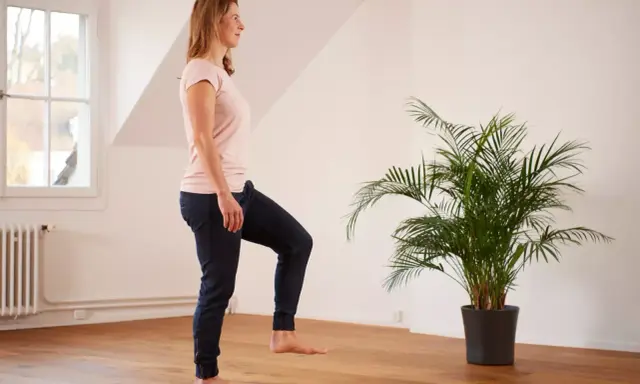 Frau übt auf einem Bein stehen im hellen Raum, neben einer grünen Palme.