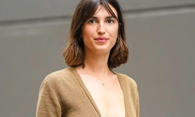 Mujer con cabello castaño ondulado, en transición de pelo corto a largo, luce un suéter marrón y un collar con cruz.