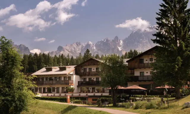 Un hotel alpino immerso nel verde, con imponenti vette innevate sullo sfondo, un perfetto scenario da montagna d'estate.
