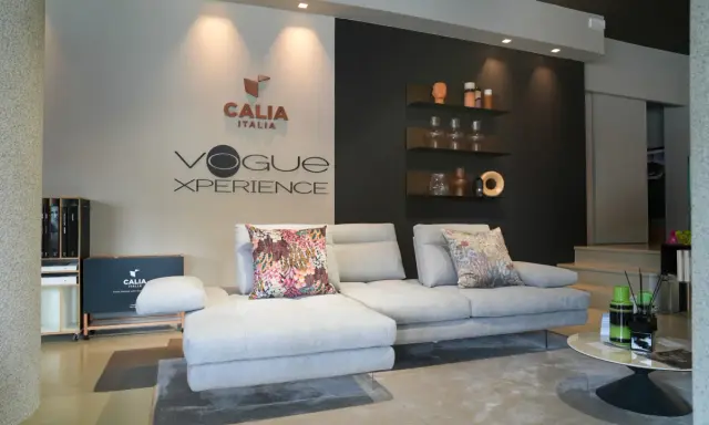Divano componibile grigio chiaro con cuscini decorativi, logo Calia Italia e "Vogue Xperience" sulla parete.