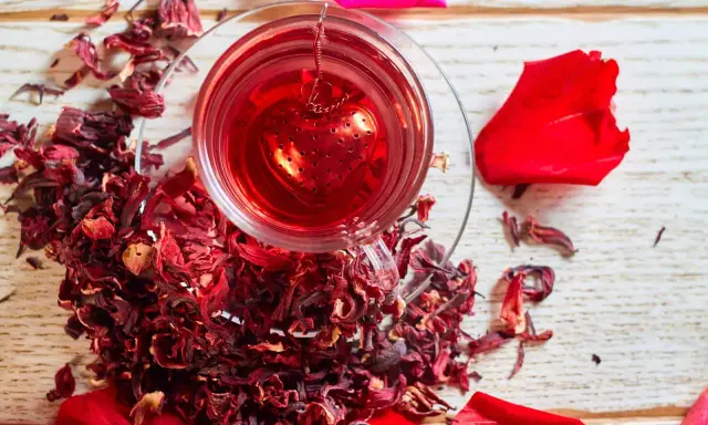 Czerwona, aromatyczna herbata z hibiskusem w szklance z zaparzaczem w kształcie serca, otoczona suszonymi kwiatami i płatkami róż.