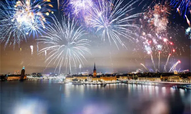 Fyrverkerier lyser upp himlen över Stockholm vid nyår. Vatten reflekterar ljusen från staden och fyrverkerierna.
