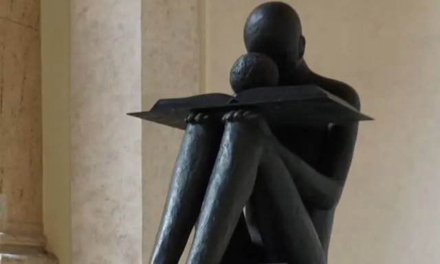 Statua di una figura che legge un libro, con un bambino appoggiato, immersi nella conoscenza a tutto spiano.