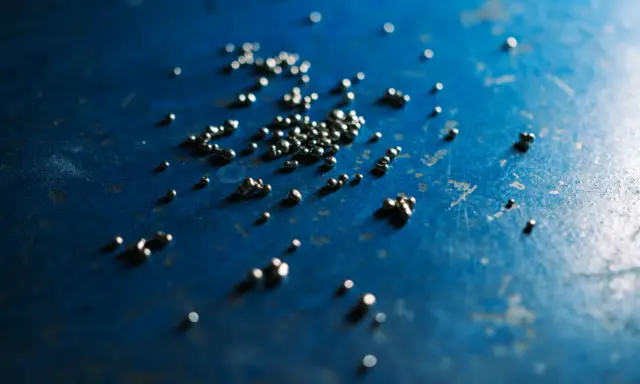 Piccole perle di argento riciclato sparse su una superficie blu scuro, con riflessi luminosi.
