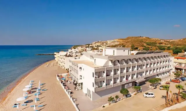 Wypoczynek na plaży w hotelu Island Maya Resort. Biały budynek hotelowy z balkonami, obok piaszczysta plaża z leżakami i parasolami, morze i wzgórza w tle.