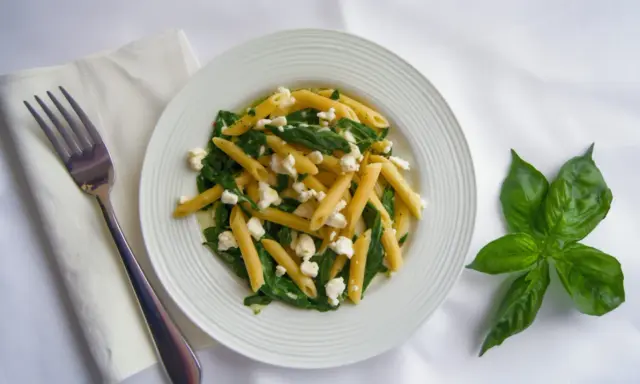Pyszny makaron penne ze szpinakiem i twarogiem, udekorowany listkami bazylii. Obok leży widelec i biała serwetka.