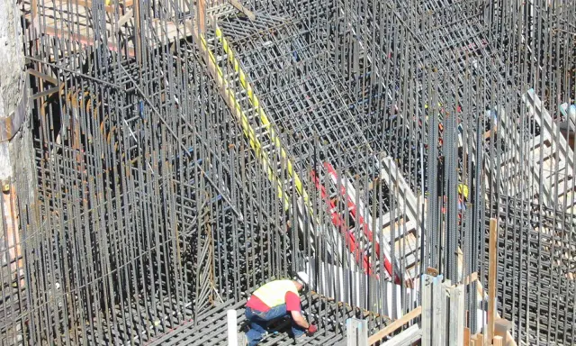 Operaio lavora su un'impalcatura di ferro per il cemento armato. La sua presenza suggerisce la cura necessaria per garantire la durata del cemento armato.