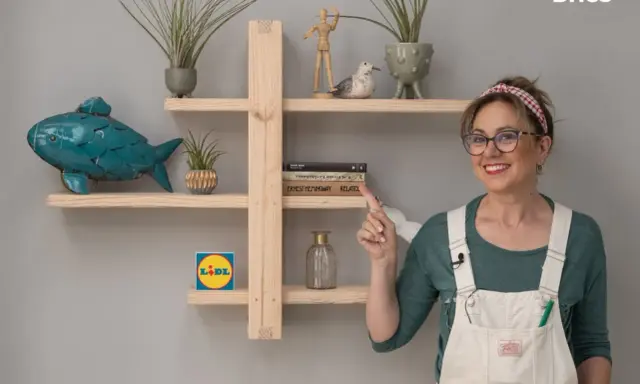 Una mujer sonriente muestra una estantería de madera, decorada con plantas, un pez azul y libros. Ideal para aprender como hacer una estantería de madera.