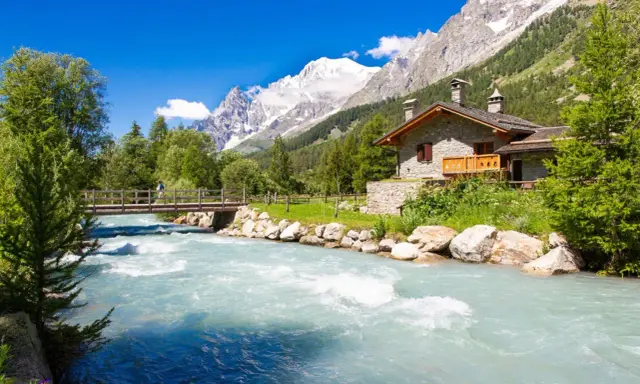 Val Ferret: un'incantevole casa in pietra alpina vicino a un fiume turchese, con un ponte e montagne innevate sullo sfondo.