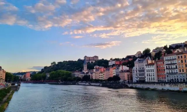 Blick auf die Stadt Lyon, Frankreich, mit dem Fluss Saône im Vordergrund und bunten Gebäuden, die sich den Hügel hinaufziehen. Ein Schloss thront über der Stadt.