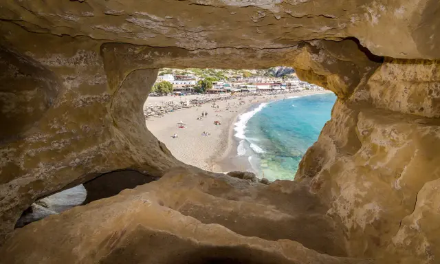 Blick durch Höhle auf Strand von Matala, Kreta. Ein sonniges Hippie-Dorf mit bunten Häusern und blauem Meer.