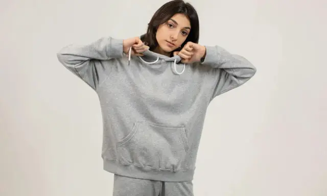 Dziewczyna w szarej bluzie oversize, z kapturem naciągniętym na głowę, pokazuje, jak nosić ten wygodny element garderoby.