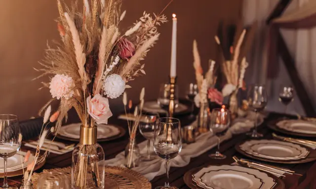 Stół weselny w stylu boho z bukietem z traw pampasowych, róż i świecami. Eleganckie nakrycie z złotymi talerzami i kieliszkami.