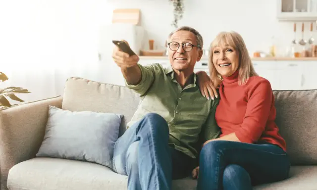 Uśmiechnięta para seniorów ogląda telewizję. Zastanawiają się, od jakiego wieku nie płaci się abonamentu RTV, ciesząc się wspólnym czasem.