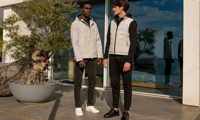 Due ragazzi con look autunnali, uno con giacca leggera, l'altro con gilet imbottito, posano vicino a un ulivo.
