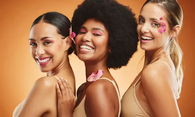 Trois femmes souriantes, un vrai "glow up traduction" avec des fleurs dans les cheveux et un maquillage éclatant.