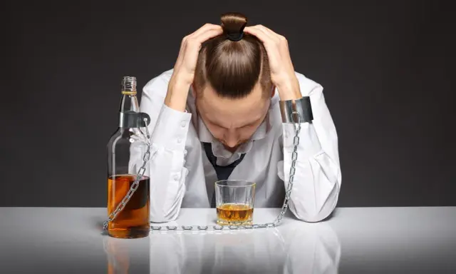 Mężczyzna z rękami na głowie, skuty łańcuchami z butelką i kieliszkiem alkoholu. To symbol walki z alkoholizmem, gdzie pomoc jest kluczowa.