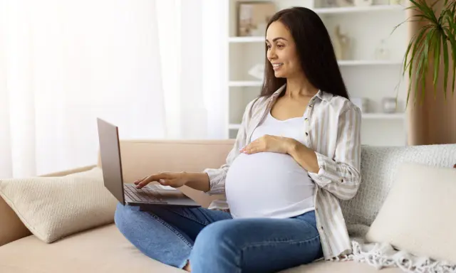 Kobieta w ciąży na kanapie szuka w internecie najdokładniejszy termin porodu, uśmiecha się.