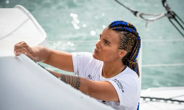 Marie Tabarly, concentrée, ajuste une voile sur son bateau. Ses tresses bleues et brunes flottent au vent.