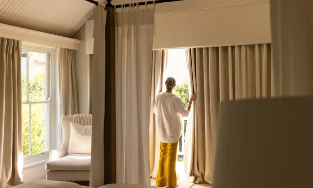 Una mujer abre cortinas para dormitorios, dejando entrar la luz. Se ven camas y un sillón junto a la ventana.