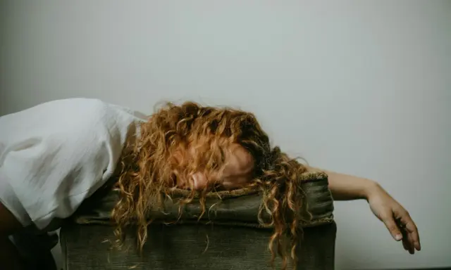 Donna con capelli rossi ricci, viso nascosto, sembra sopraffatta, forse un attacco di panico o uno svenimento.
